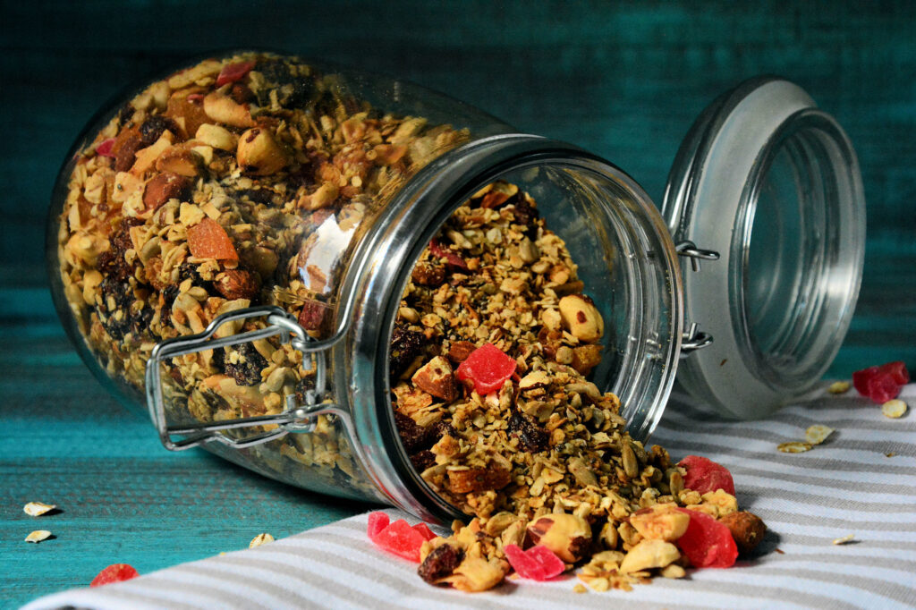 Jar tipped over, spilling granola with oats, nuts and dried fruit onto a teal surface and striped cloth in the foreground